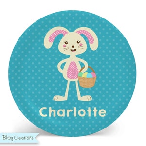 May include: A turquoise blue round plate with white polka dots. A cartoon bunny with pink ears and a pink and white polka dot dress holds a basket of colorful Easter eggs. The plate has the name "Charlotte" written in a cream color.