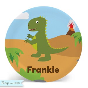 May include: A round, blue and brown plate with a green dinosaur, a volcano, and palm trees. The plate has the name "Frankie" written on it.
