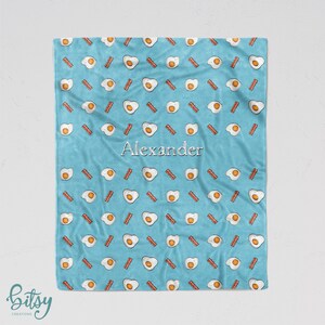 May include: A light blue fleece blanket with a repeating pattern of white eggs and brown bacon strips. The name "Alexander" is printed in white in the center of the blanket.