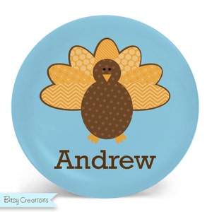 May include: A blue personalized plate with a brown and orange turkey design. The turkey has a brown body with white polka dots, a yellow beak, and orange and yellow patterned feathers. The name "Andrew" is printed in brown letters below the turkey.