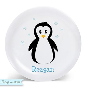 May include: White ceramic plate with a cartoon penguin illustration and the name "Reagan" in blue text. The penguin is black and white with a yellow beak and blue snowflakes are scattered around the penguin.