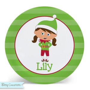 May include: A green and white striped plate with a red border. The center of the plate features a cartoon elf girl with brown hair, wearing a green and white hat, a green shirt, a brown belt, red pants, and brown shoes. The elf girl's name, "Lily", is written in red letters below her.
