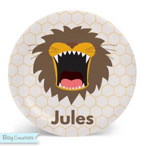May include: A round white plate with a geometric pattern in gold. The center of the plate features a cartoon lion with an open mouth and sharp teeth. The name "Jules" is printed below the lion.