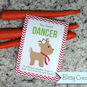 May include: A red and white chevron patterned card with the text "You found DANCER" and a cartoon reindeer illustration. The text below the reindeer says "A different reindeer loves to read. You'll find him with books!" The card is sitting on top of three orange carrots.