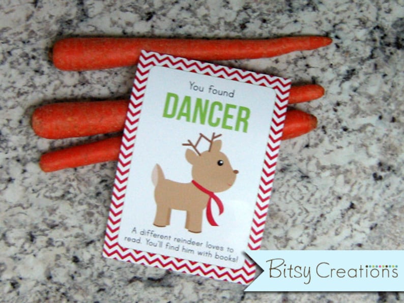 May include: A red and white chevron patterned card with the text "You found DANCER" and a cartoon reindeer illustration. The text below the reindeer says "A different reindeer loves to read. You'll find him with books!" The card is sitting on top of three orange carrots.