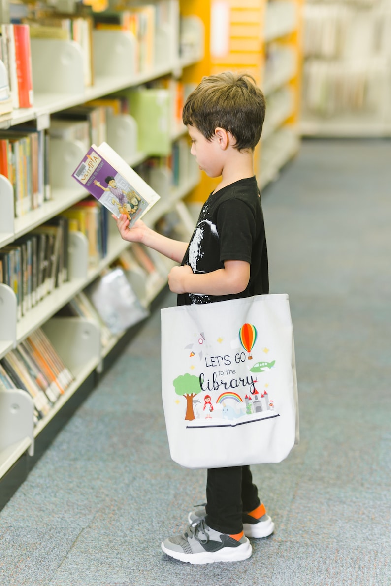 May include: A young person in a black shirt and black pants is holding a book and a white tote bag with the text "Let's go to the library" printed on it. The bag has a colorful design with a rainbow, a hot air balloon, a castle, and a tree.