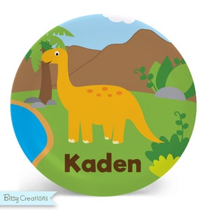 May include: A green and yellow dinosaur themed plate with a yellow dinosaur and the name "Kaden" printed on it.