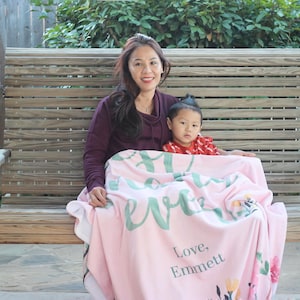 Pu&ograve; includere: Una donna e un bambino piccolo sono seduti su un'altalena di legno. Sono entrambi avvolti in una coperta rosa con un motivo floreale verde e le parole "Sei tutto per me" e "Con amore, Emmett".