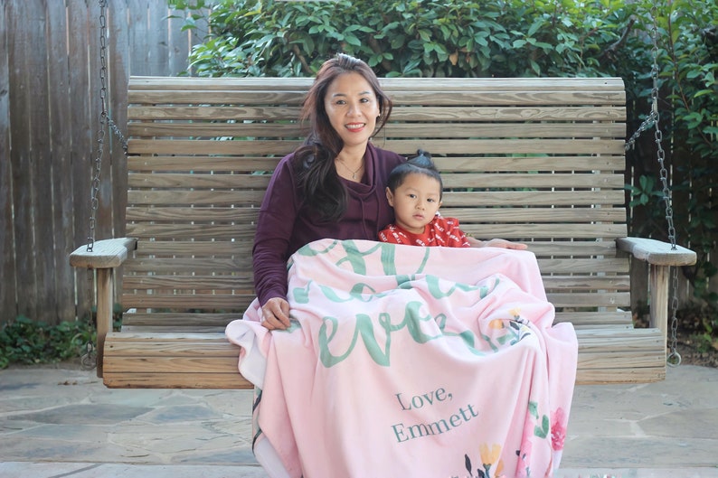 Pu&ograve; includere: Una donna e un bambino piccolo sono seduti su un'altalena di legno. Sono entrambi avvolti in una coperta rosa con un motivo floreale verde e le parole "Sei tutto per me" e "Con amore, Emmett".