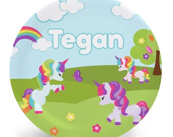 May include: A personalized plate with a blue sky, green grass, and two colorful unicorns. The plate has a rainbow and the name "Tegan" written in white.