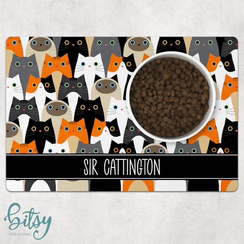 Pode incluir: Uma tigela branca para comida de gato com ra&ccedil;&atilde;o marrom dentro, sentada em um tapete para animais de estima&ccedil;&atilde;o com tema de gato preto e branco com o texto "SIR CATTINGTON" em letras brancas. O tapete apresenta um padr&atilde;o repetitivo de gatos coloridos com diferentes cores de olhos.