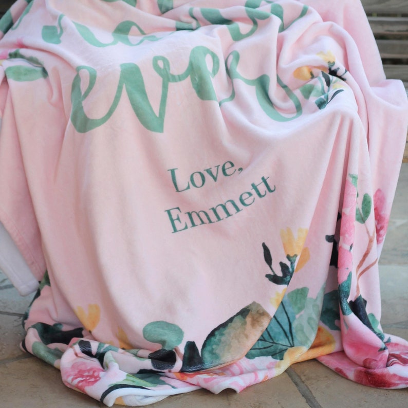 Pu&ograve; includere: Una coperta in pile rosa con un motivo floreale e il testo "Love, Emmett" in verde.