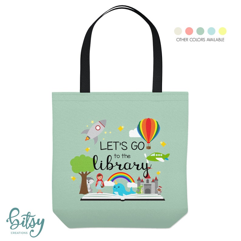 May include: A green canvas tote bag with black handles. The bag features a colorful illustration of a book with a rocket, hot air balloon, airplane, and a rainbow. The text "Let's go to the library" is printed on the bag.