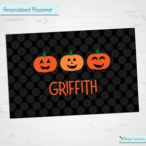 May include: Black and orange Halloween-themed placemat with three smiling jack-o'-lantern faces and the name "GRIFFITH" in orange text.