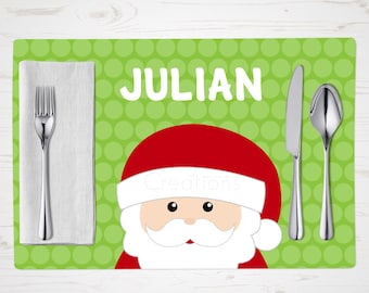 May include: A personalized Christmas placemat featuring a Santa Claus design with a red hat and white beard. The name "JULIAN" is printed in white block letters on a green polka-dot background. Silverware and a white napkin are placed on the left.