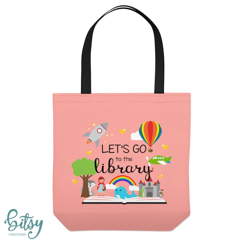 May include: Pink canvas tote bag with black handles. The bag features a colorful illustration of a book with a rocket, hot air balloon, airplane, and a rainbow. The text "Let's Go to the Library" is printed on the bag.