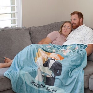 May include: A couple cuddles on a couch under a blue fleece blanket with a personalized wedding design featuring a cartoon bride and groom. The blanket has the names "Bridgette" and "Jason" printed on it.