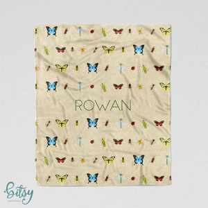 May include: A cream-colored fleece blanket with a repeating pattern of butterflies, ladybugs, dragonflies, and bees. The blanket has the name "ROWAN" printed in green letters in the center.
