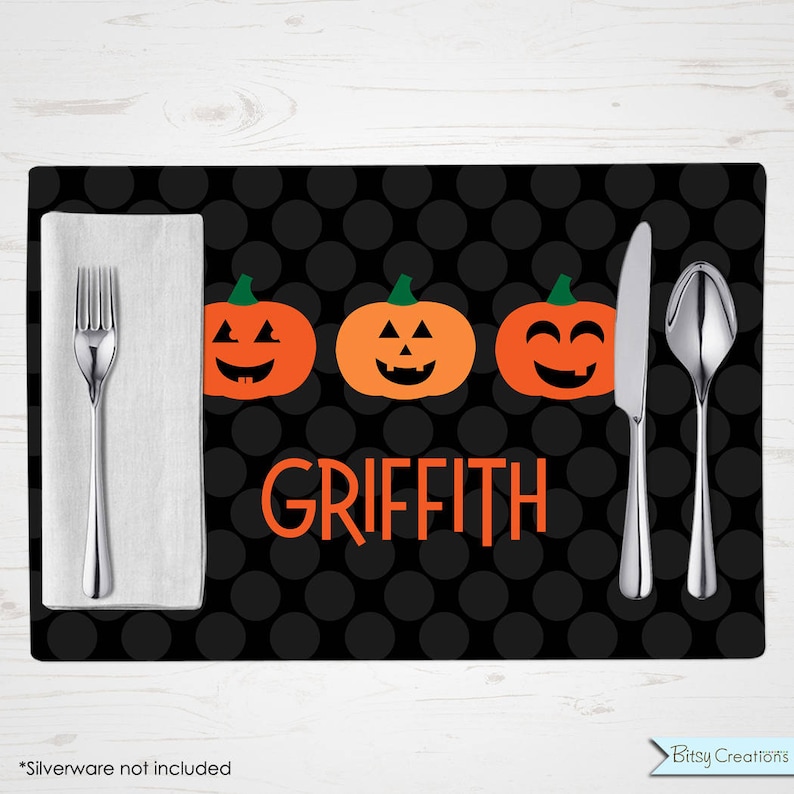 May include: Black and orange Halloween placemat with a black polka dot pattern. The placemat features three orange jack-o'-lantern faces with smiling expressions. The name "Griffith" is printed in orange letters on the placemat.