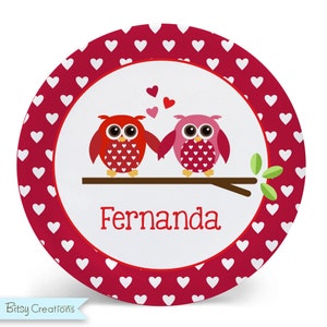 May include: A white round plate with a red border and white hearts. The center of the plate has two owls holding hands, one red and one pink, with hearts floating above them. The text "Fernanda" is written below the owls.