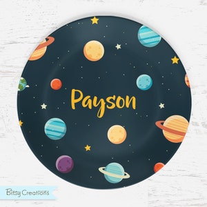 May include: A dark blue round plate with a space theme. The plate is decorated with colorful planets, stars, and the name "Payson".