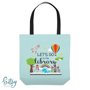 May include: A light blue tote bag with black handles. The bag has a colorful illustration of a book, a rocket, a hot air balloon, a plane, a rainbow, and a narwhal. The text "Let's go to the library" is printed on the bag.