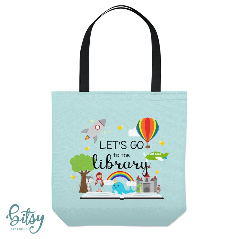 May include: A light blue tote bag with black handles. The bag has a colorful illustration of a book, a rocket, a hot air balloon, a plane, a rainbow, and a narwhal. The text "Let's go to the library" is printed on the bag.