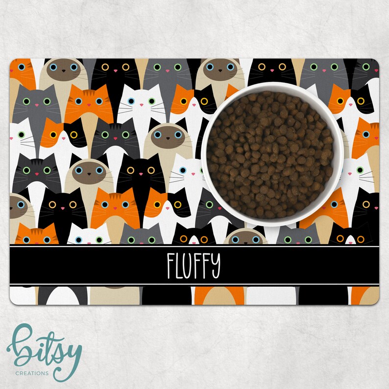 Pode incluir: Um tapete para tigelas de animais de estima&ccedil;&atilde;o com tema de gato preto e branco com uma tigela branca e comida para animais de estima&ccedil;&atilde;o marrom. O tapete tem uma borda preta com o nome "FLUFFY" impresso em branco.