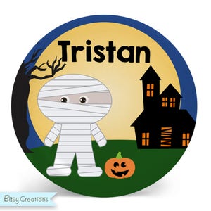 May include: A round, blue and yellow Halloween-themed personalized plate with the name "Tristan" printed on it. A cartoon mummy is standing next to a jack-o-lantern in front of a black silhouette of a haunted house.