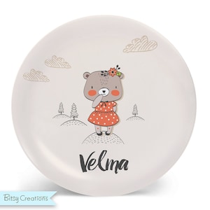 May include: A white ceramic plate with a brown bear wearing a red polka dot dress and a flower crown. The bear is standing on a hill with trees in the background. The plate has the name "Velma" written on it in black.