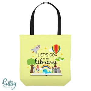 May include: A yellow tote bag with black handles. The bag has a colorful illustration of a book, a rocket, a hot air balloon, a plane, a unicorn, a castle, and a girl in a red dress. The text on the bag reads "Let's go to the library".