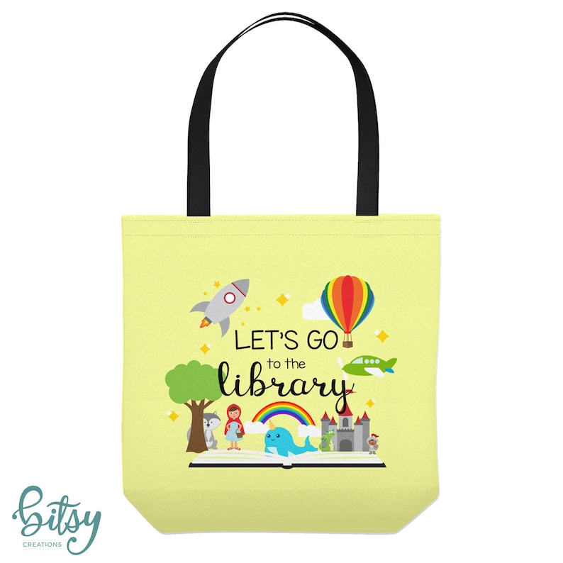May include: A yellow tote bag with black handles. The bag has a colorful illustration of a book, a rocket, a hot air balloon, a plane, a unicorn, a castle, and a girl in a red dress. The text on the bag reads "Let's go to the library".