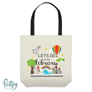 May include: A white canvas tote bag with black handles. The bag features a colorful illustration of a book with a rainbow, a rocket, a hot air balloon, a plane, and a castle. The text "Let's go to the library" is printed in black on the bag.
