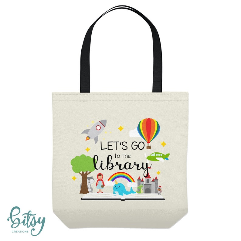 May include: A white canvas tote bag with black handles. The bag features a colorful illustration of a book with a rainbow, a rocket, a hot air balloon, a plane, and a castle. The text "Let's go to the library" is printed in black on the bag.