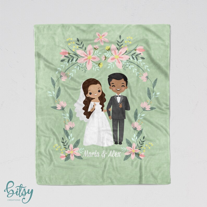 May include: A green fleece blanket with a floral wreath design. The wreath features pink flowers and green leaves. Inside the wreath is a cartoon illustration of a bride and groom. The bride is wearing a white dress and the groom is wearing a black suit. The text "Maria & Alex" is written below the couple.