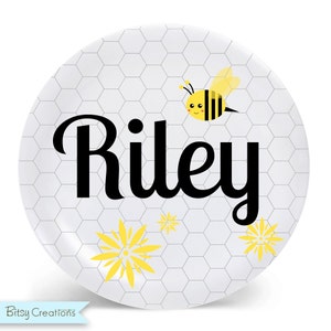 May include: A white ceramic plate with a honeycomb pattern, a yellow and black bee, and the name "Riley" in black cursive. The plate has yellow flower accents.