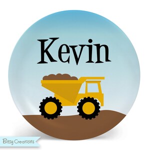 May include: A yellow toy dump truck with brown dirt in the bed. The truck is on a brown surface with a blue sky in the background. The name "Kevin" is written in black letters above the truck.