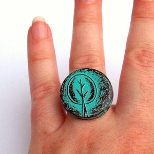 May include: A turquoise blue ring with a distressed finish features a raised, black silhouette of a tree with branches. The ring is worn on a finger.