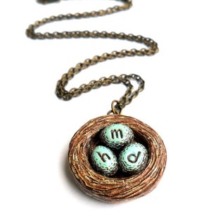 May include: A necklace with a brown bird's nest pendant. The nest contains three blue eggs with the letters 'M', 'H', and 'D' inscribed on them.