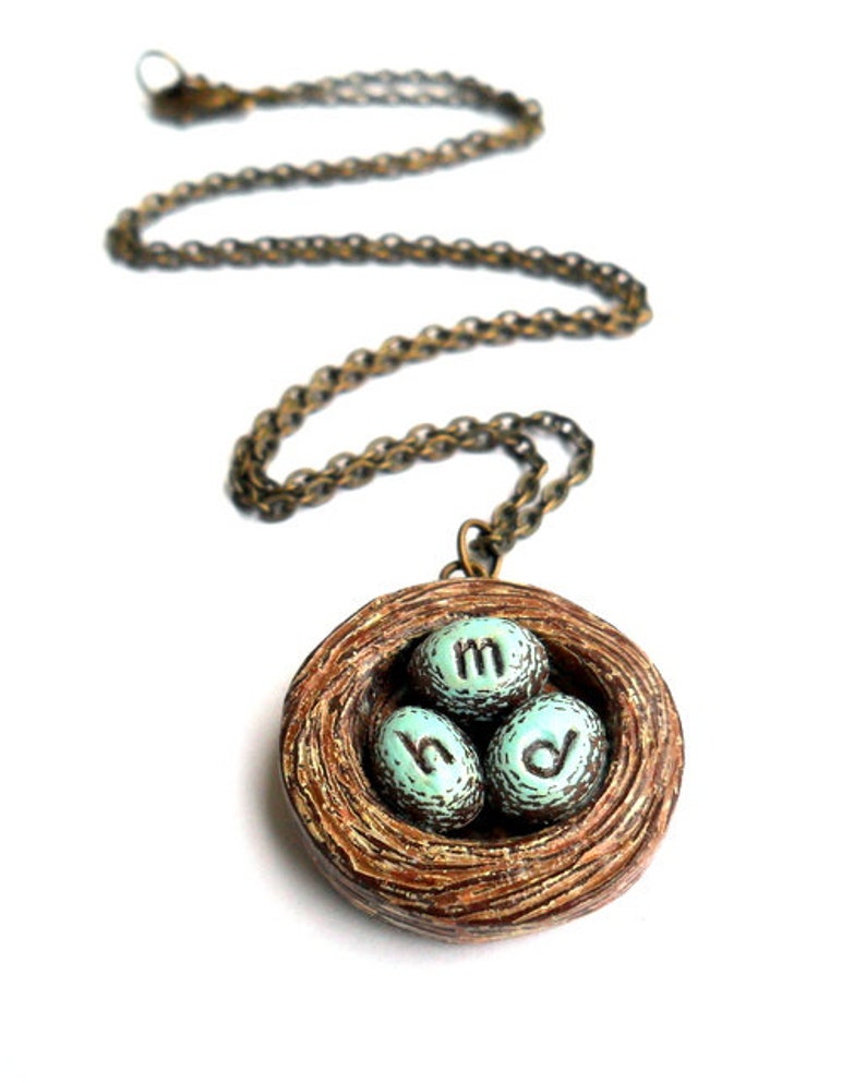 May include: A necklace with a brown bird's nest pendant. The nest contains three blue eggs with the letters 'M', 'H', and 'D' inscribed on them.