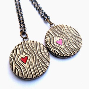 May include: Two round pendant necklaces with a wood grain design and a heart-shaped center. One heart is red, the other is pink. The pendants hang from a dark chain.