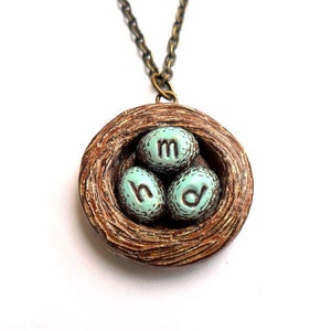 May include: A brown bird's nest necklace with three blue eggs that have the letters 'm', 'h', and 'd' on them.