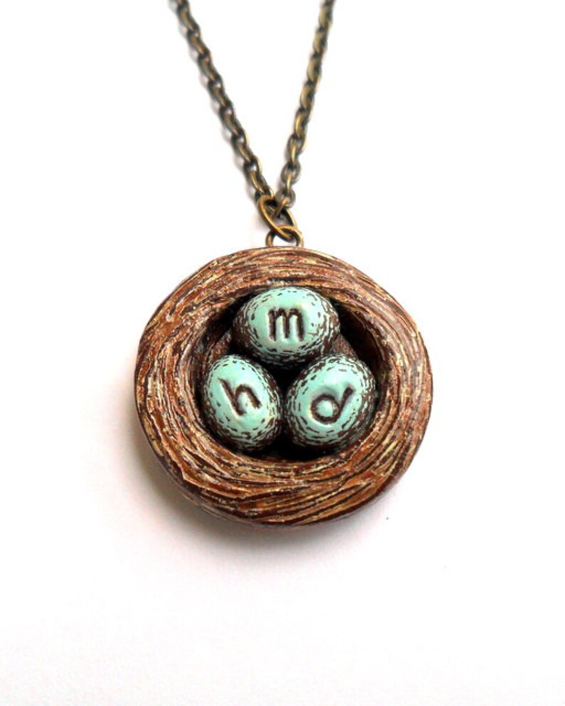 May include: A brown bird's nest necklace with three blue eggs that have the letters 'm', 'h', and 'd' on them.