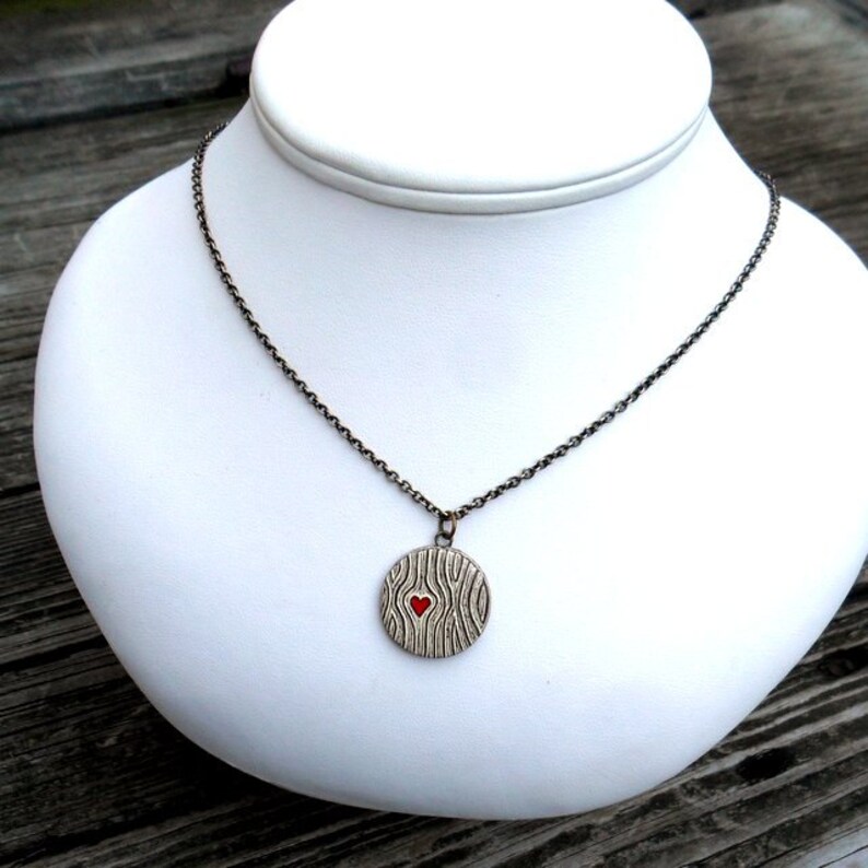 May include: A silver-toned necklace with a round pendant featuring a wood grain design and a small red heart in the center. The necklace is displayed on a white bust against a blurred wooden background.