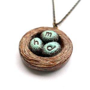 May include: A brown ceramic bird's nest necklace with three teal blue eggs that have the letters 'h', 'o', and 'm' on them.