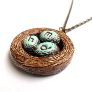 May include: A brown ceramic bird's nest necklace with three blue eggs that have the letters 'H', 'M', and 'O' painted on them.