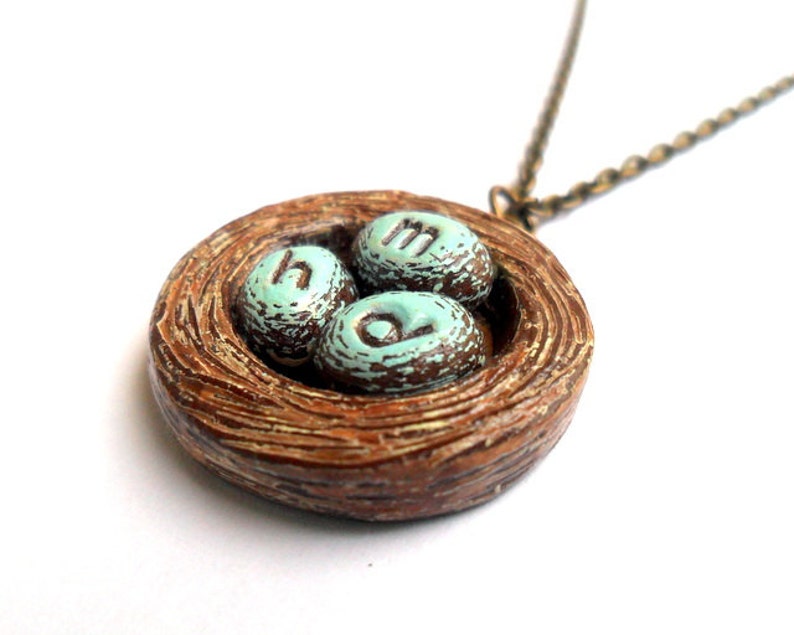 May include: A brown ceramic bird's nest necklace with three blue eggs that have the letters 'H', 'M', and 'O' painted on them.