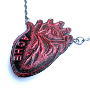 May include: A red and black heart-shaped pendant necklace. The pendant has the word "ACHE" on it. The necklace is made of metal and has a chain. The heart pendant has a textured surface.