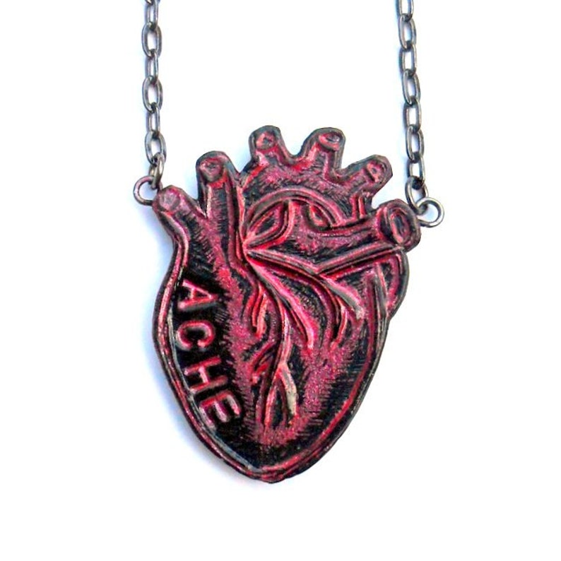 May include: A red and black heart pendant necklace. The pendant is shaped like a human heart with the word "ACHE" on the side. The necklace chain is a dark silver color.