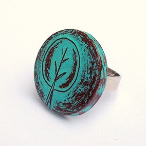 May include: A turquoise blue ring with a brown and black tree design. The ring is made of metal and has a distressed look.